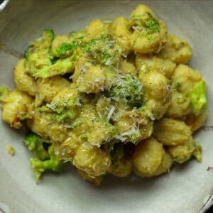 A bowl of creamy broccoli gnocchi mixed with chopped broccoli and topped with grated cheese, all coated in a light greenish-brown sauce. The dish is served in a rustic, light-colored ceramic bowl.